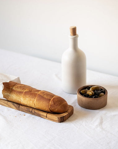 An olive oil bottle and a loaf of bread on a wooden board, with a small wooden bowl of olives beside it, all placed on a white surface.