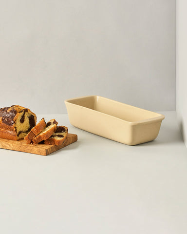 A white enameled stoneware cake and bread pan on a beige background, with two red apples placed on top of it to the side.