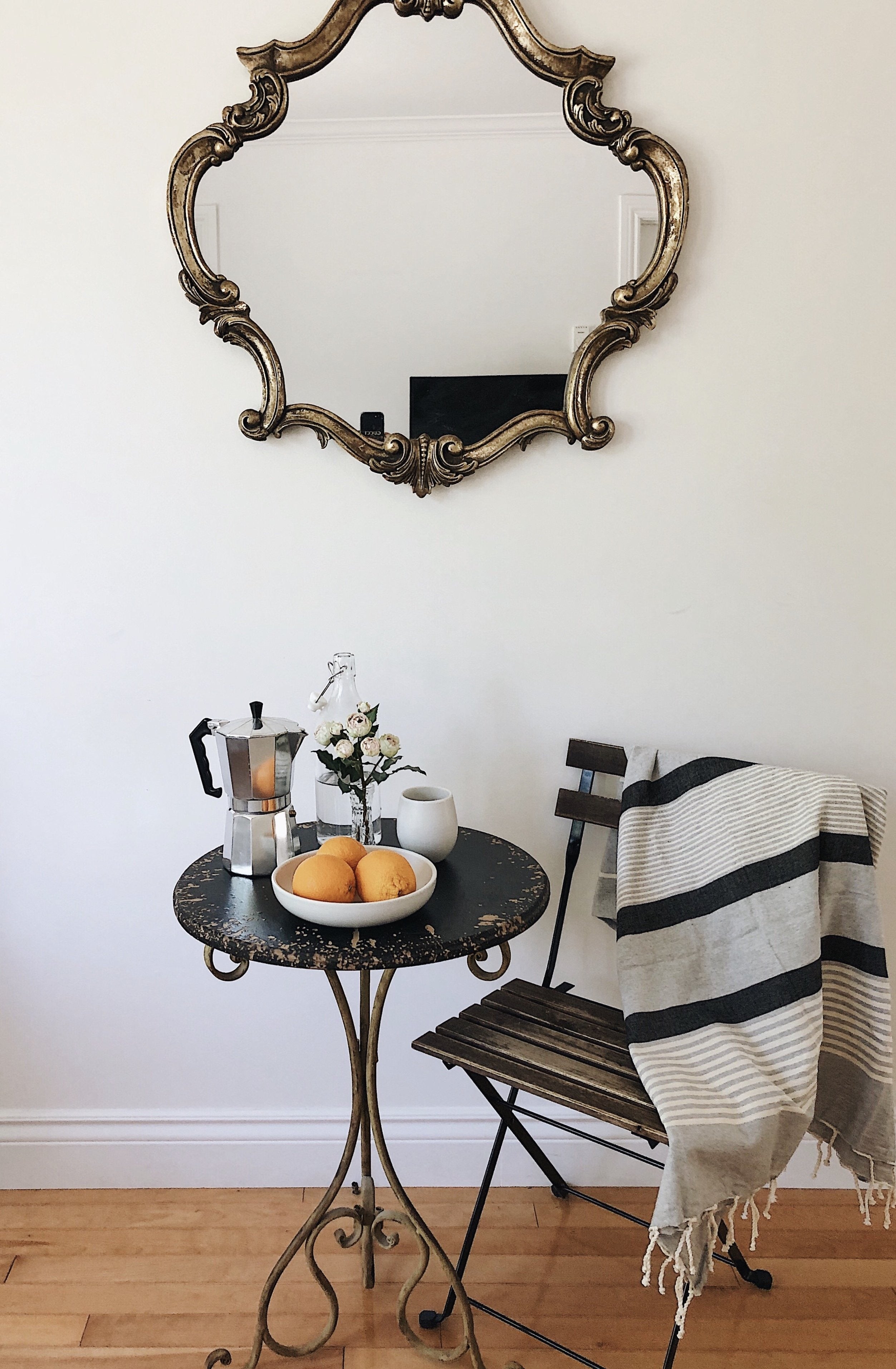 A gray fouta towel with black stripes draped over the back of a chair. Next to it is a set dining table with fruits, and a decorative design is visible on the wall in the background, creating a cozy and styled scene.
