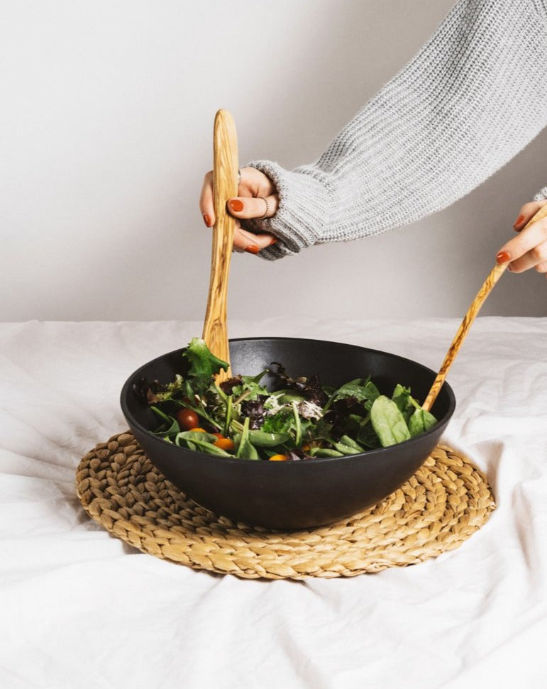 A person's hands using olive wood serving forks to serve salad from a black stoneware bowl, which is placed on a beige mat.