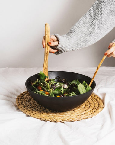 A person's hands using olive wood serving forks to serve salad from a black stoneware bowl, which is placed on a beige mat.