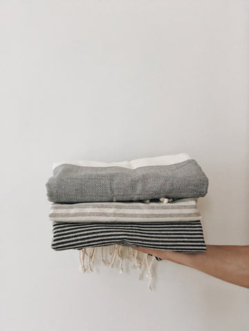 A gray fouta towel with black stripes, neatly folded and held up by hand to showcase its texture, pattern, and lightweight fabric.

