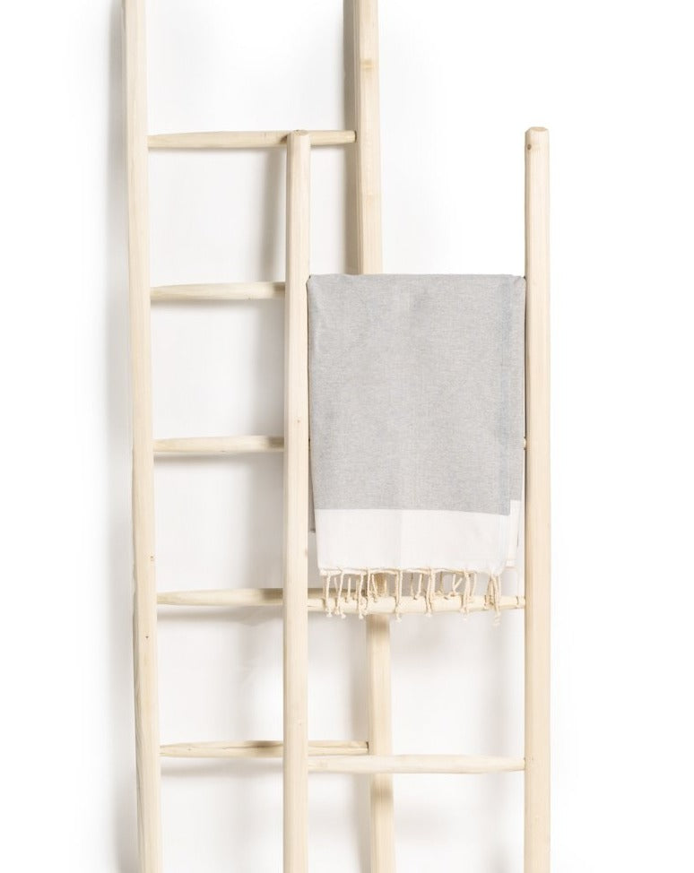 Two wooden ladders set against a white background, with a gray Fouta towel draped over one of them, showcasing its texture and striped design in a minimalist setting.