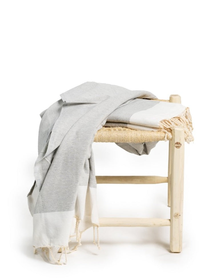 A gray Fouta towel draped over a wooden chair, set against a plain white background to highlight the towel’s texture and design.

