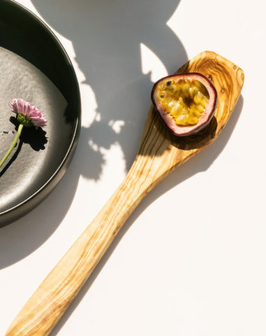 Olive wood spoon holding an unfamiliar food item, placed beside a half black bowl containing a picked flower.