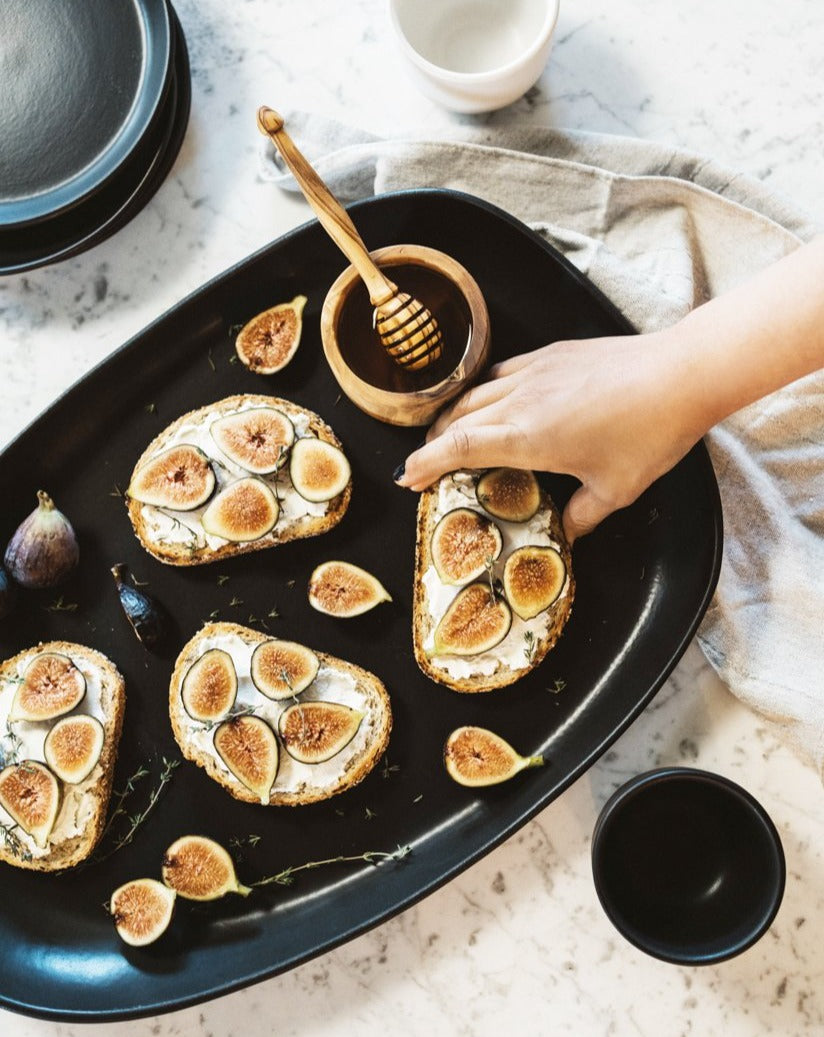 A person's hand reaching for food arranged on a black stoneware serving platter on a table, with a honey dipper and figs visible on the platter.