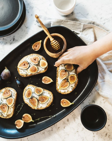 A person's hand reaching for food arranged on a black stoneware serving platter on a table, with a honey dipper and figs visible on the platter.