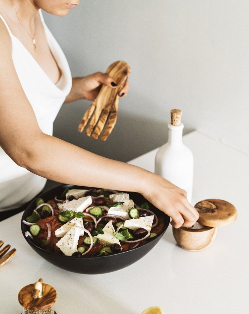 flat olive wood serving fork being used by a person, with their face partially out of frame, over a bowl of salad on a table alongside a cork-topped dispenser.