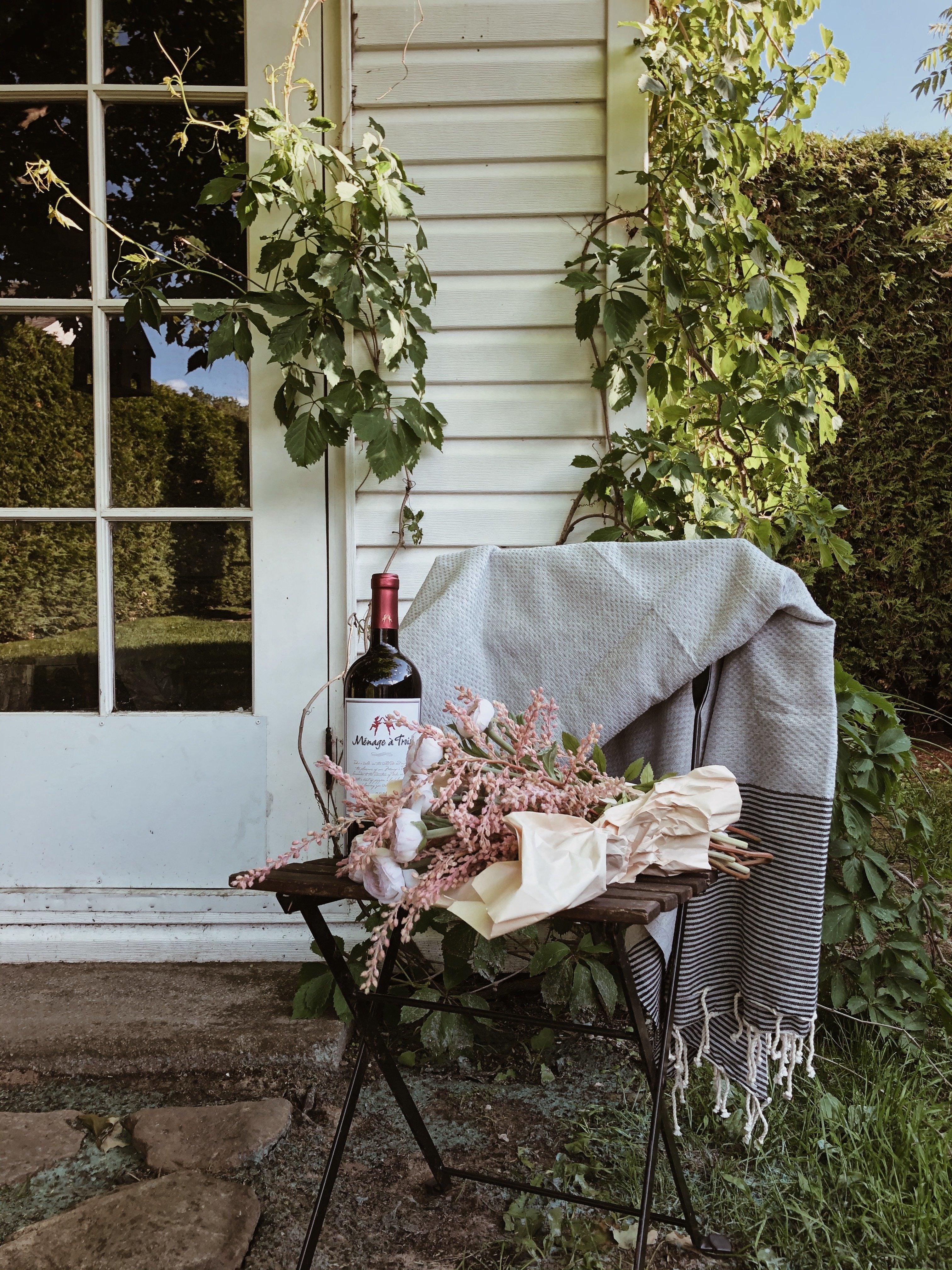 A Fouta towel is displayed outdoors, draped over a chair next to a glass door, with a bottle and some decorative flowers on the table, set against a backdrop of greenery.