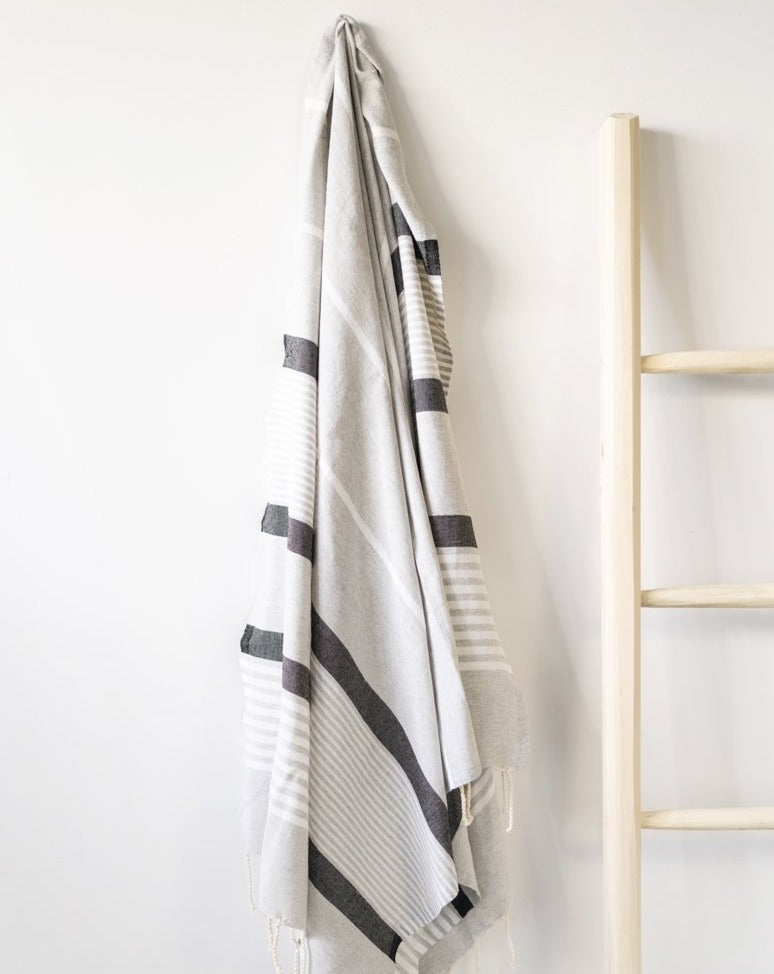 A gray fouta towel with black stripes is hanging on a wall next to a wooden ladder.
