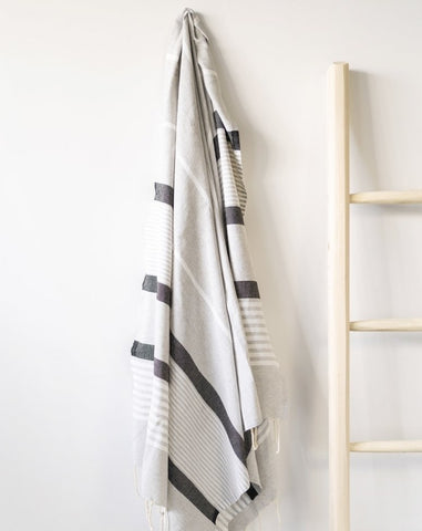 A gray fouta towel with black stripes is hanging on a wall next to a wooden ladder.
