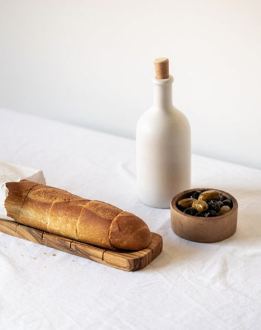 Olive wood bread slicing board with bread on top, accompanied by a snack bowl with contents and a cork-topped dispenser, set against a white background.