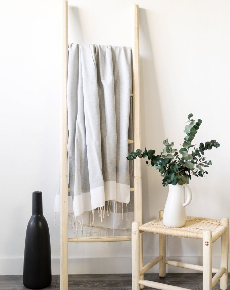  A gray Fouta towel hanging on a wooden ladder against a white wall. Beside it is a wooden chair with a vase of greenery placed on the seat, and a dispenser is positioned nearby, creating a natural and calming interior scene.

