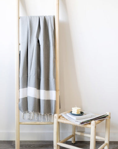 A gray fouta towel with white stripes, draped over a wooden ladder, next to a small wooden table with a book and a candle.