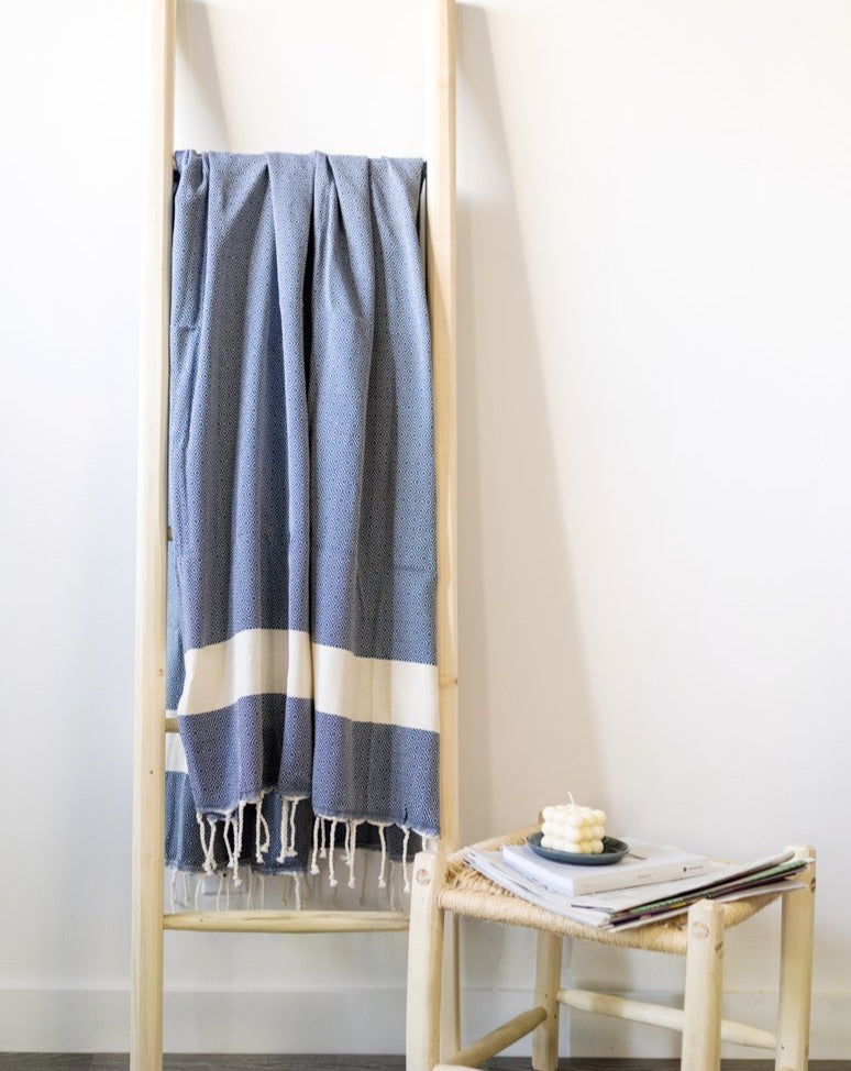A sky blue Fouta towel with a diamond pattern and a white stripe, displayed on a wooden ladder, used as a throw.
