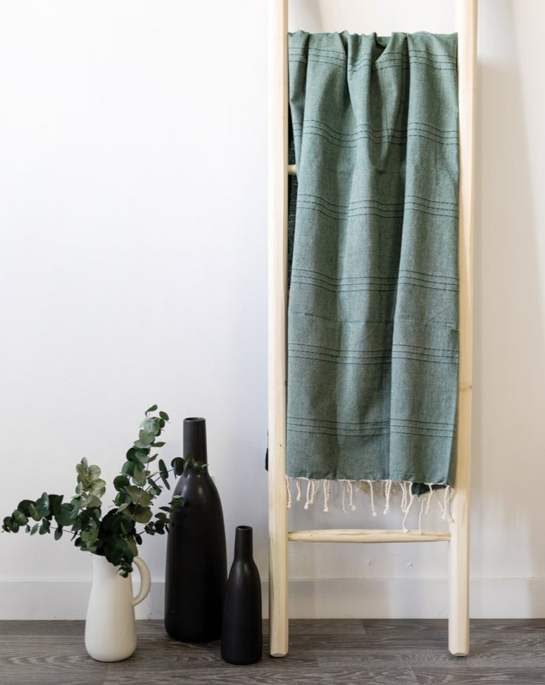 A green fouta towel hanging on a wooden ladder with a vase of flowers and bottles beside it.