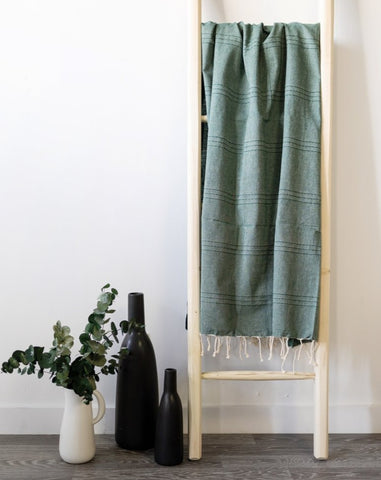 A green fouta towel hanging on a wooden ladder with a vase of flowers and bottles beside it.